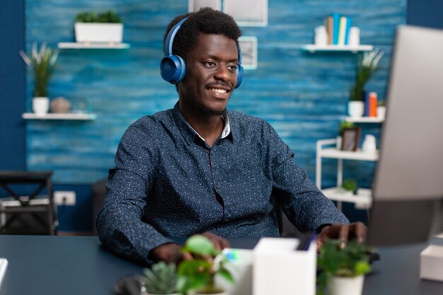 Homem afro-americano sorridente autêntico usando laptop e fones de ouvido para trabalhar em casa e aprender freel ...