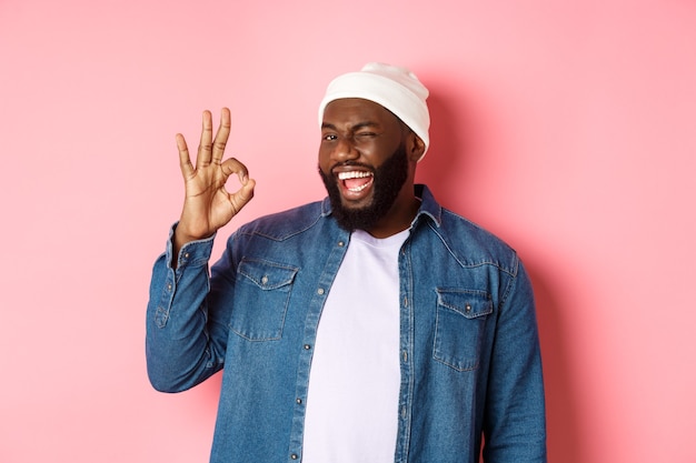 Homem afro-americano sorridente feliz mostrando sinal de tudo bem, aprovar e elogiar a boa oferta, em pé sobre o fundo rosa.