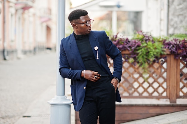 Homem afro-americano surpreendentemente vestindo blazer azul com broche de gola alta preta e óculos posando na rua Cara negro na moda
