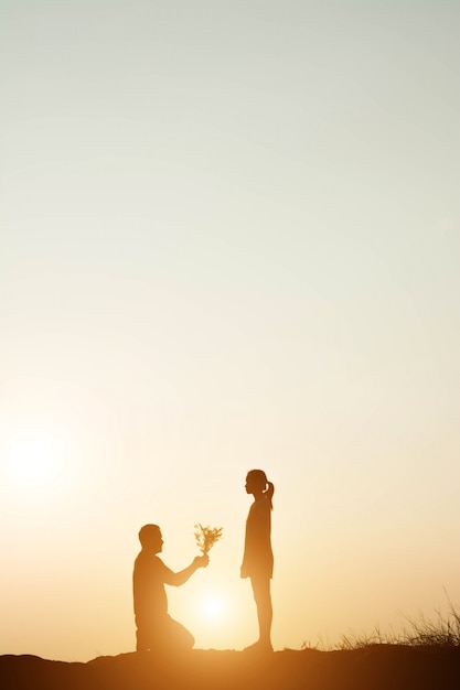 Homem ajoelhado ao pôr do sol dá o ramalhete a uma mulher