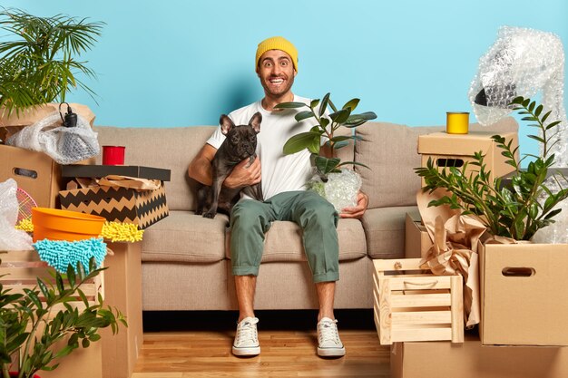 Homem alegre abraça cachorro e maconha com planta de casa, sentado na sala de estar no sofá