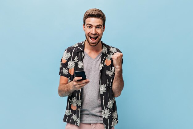 Homem alegre com olhos azuis em roupas legais da moda olhando para a câmera se alegra e posando com smartphone em pano de fundo isolado