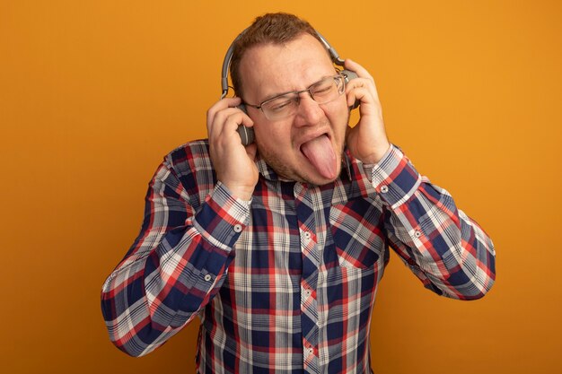 Homem alegre de óculos e camisa xadrez com fones de ouvido e a língua para fora em pé sobre uma parede laranja