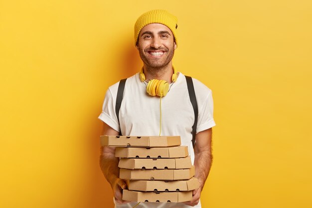 Homem alegre e alegre em trajes casuais, segura pilha de caixas de papelão com pizza, tem expressão amigável, usa fones de ouvido para ouvir faixa de áudio, entrega junk food, demonstra bom serviço