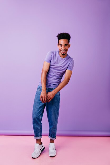 Homem alegre e emocional com aparência engraçada de penteado. Negro interessado em uma camiseta roxa posando com um sorriso tímido