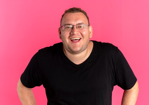 Homem alegre e obeso de óculos, vestindo uma camiseta preta com um grande sorriso no rosto em pé sobre uma parede rosa