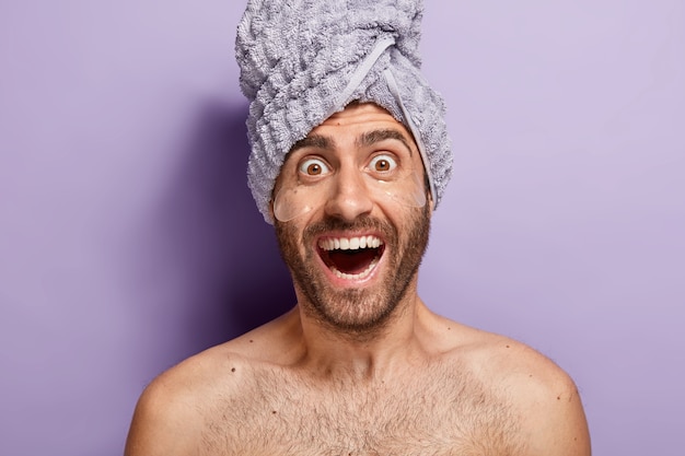 Homem alegre e surpreso com almofadas sob os olhos, sem camisa contra um fundo roxo, usa uma toalha na cabeça, se preocupa com a pele do rosto