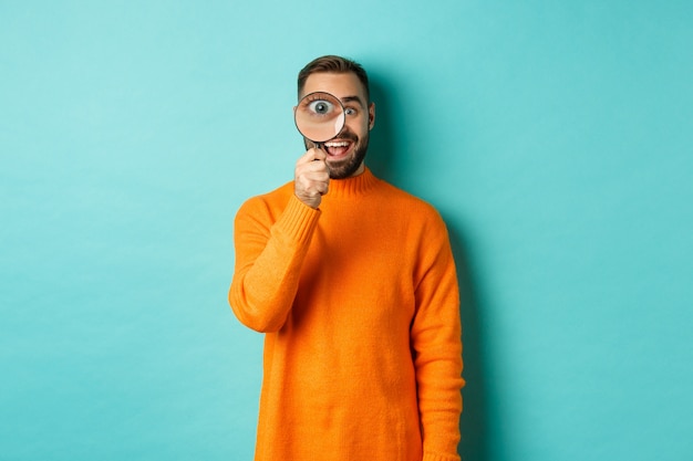 Homem alegre procurando por algo, olhando pela lupa e sorrindo feliz, encostado na parede turquesa