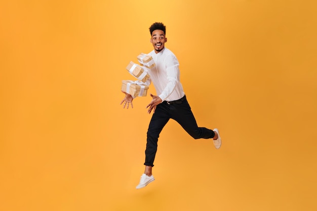 Foto grátis homem alegre pula em fundo laranja e vomita presentes homem de cabelos escuros de camisa branca e calças pretas posando alegremente em isolado