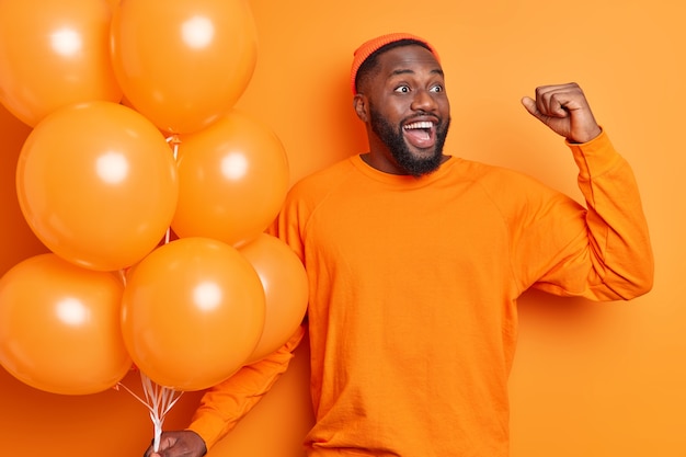 Foto grátis homem alegre se sente como se o vencedor levantasse o braço, cerrasse o punho, olhasse alegremente para o lado, celebra a obtenção de uma nova posição no trabalho estando em uma festa corporativa segurando um monte de suportes de balões inflados