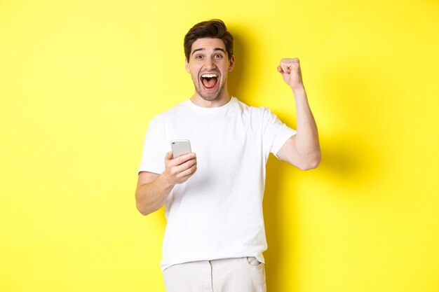 Homem alegre vencendo no smartphone, levantando a mão e segurando o celular, atingir a meta do aplicativo, em pé sobre um fundo amarelo