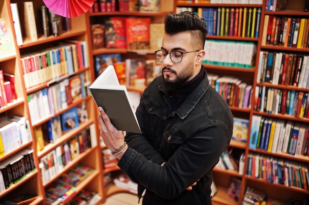 Homem alto estudante árabe inteligente usa jaqueta jeans preta e óculos na biblioteca com livro nas mãos