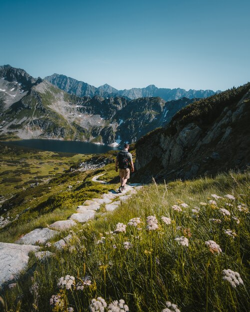 Homem andando em um caminho de pedra, rodeado por montanhas, plantas e um lago