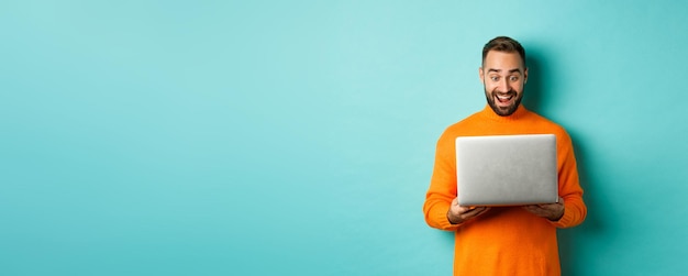 Foto grátis homem animado com suéter laranja trabalhando no laptop olhando para a tela do computador espantado em pé sobre a luz