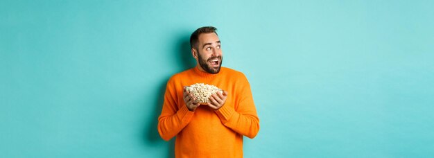 Homem animado e feliz assistindo tv e segurando uma tigela de pipoca olhando para a esquerda e sorrindo satisfeito