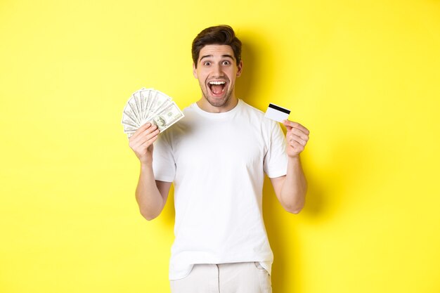 Homem animado, pronto para fazer compras de sexta-feira negra, segurando dinheiro e cartão de crédito, em pé sobre fundo amarelo.