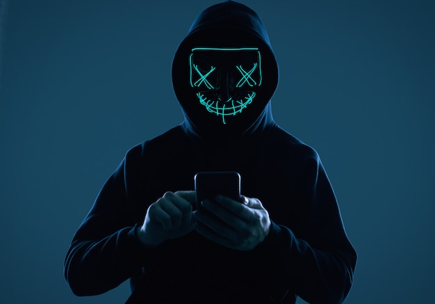 Homem anônimo em um capuz preto e máscara de néon invadindo um smartphone