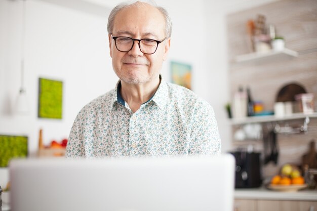 Homem aposentado sorrindo enquanto assiste a um filme no laptop. vida diária do homem sênior na cozinha durante o café da manhã, usando laptop segurando uma xícara de café. idoso aposentado trabalhando em casa, telecommu