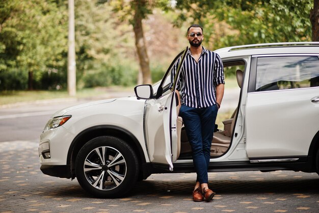 Homem árabe bem sucedido usa camisa listrada e óculos de sol posam perto de seu carro suv branco Homens árabes estilosos em transporte