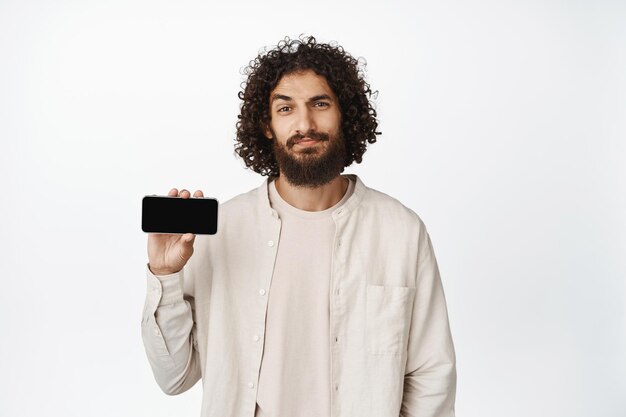Homem árabe bonito sorridente mostrando a interface do aplicativo de tela do smartphone horizontal em roupas casuais sobre fundo branco