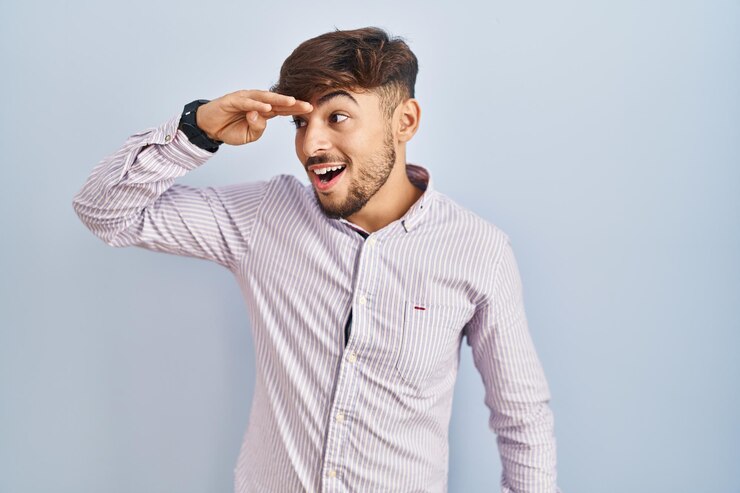 Foto grátis homem árabe com barba em pé sobre fundo azul muito feliz e sorridente olhando para longe com a mão sobre a cabeça. conceito de pesquisa.