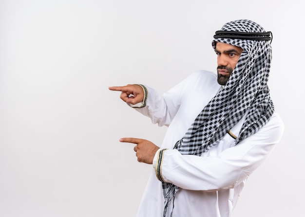 Homem árabe com roupa tradicional olhando para o lado com o rosto sério apontando com os dedos para o lado em pé sobre uma parede branca