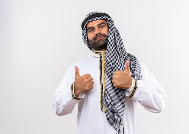 Foto grátis homem árabe com roupa tradicional sorrindo e mostrando os polegares em pé sobre uma parede branca