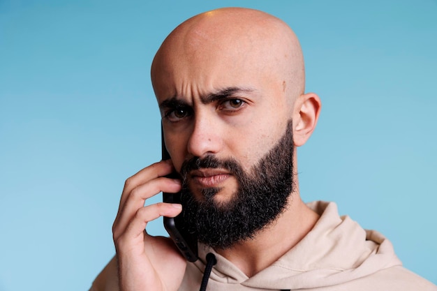 Foto grátis homem árabe confuso atendendo chamada de smartphone e retrato de sobrancelhas carrancudas. jovem barbudo careca falando no celular e olhando para a câmera com expressão incerta