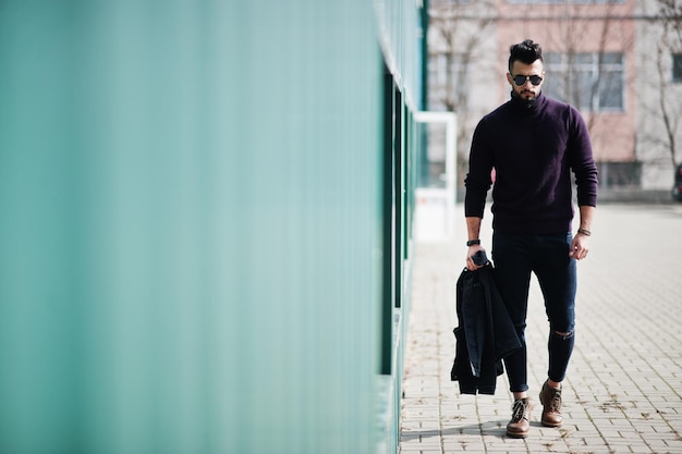 Homem árabe de barba rica de moda usa na gola alta e óculos de sol andando contra a parede verde com jaqueta na mão Cara de modelo árabe elegante, bem-sucedido e elegante