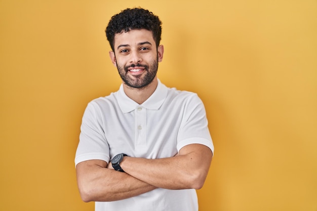 Homem árabe de pé sobre o rosto feliz de fundo amarelo sorrindo com os braços cruzados, olhando para a câmera. pessoa positiva.