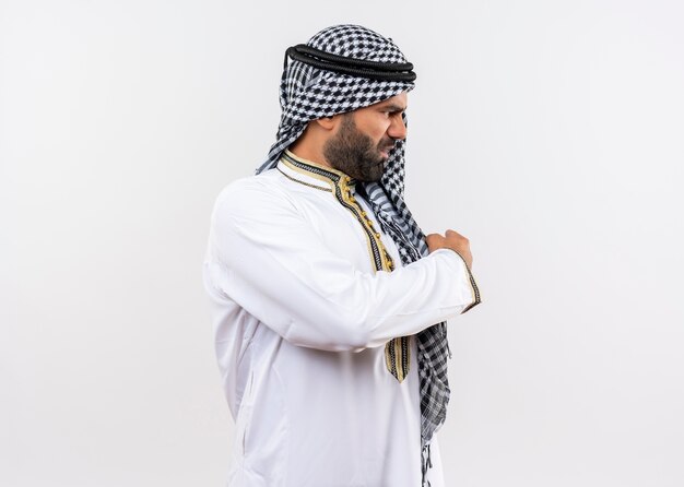 Homem árabe descontente com roupas tradicionais olhando para o lado e apontando para trás com o dedo em pé sobre uma parede branca