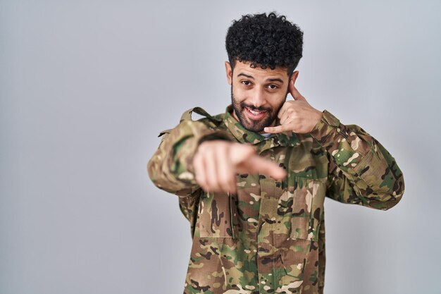 Homem árabe vestindo uniforme do exército de camuflagem sorrindo fazendo falar no gesto de telefone e apontando para você. liga para mim.