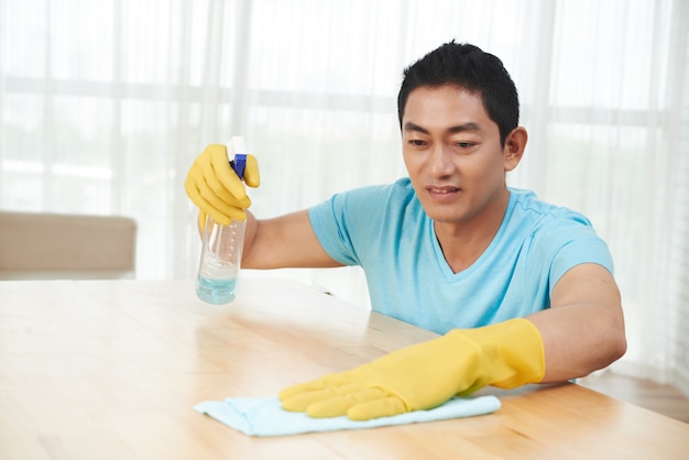 Homem asiático em luvas de borracha, mesa de pulverização e limpeza com pano em casa