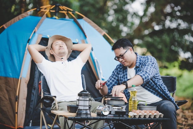 Homem asiático relaxado usando smartphone e esperando por seu amigo cozinhando com fundo de tenda no acampamento cozinhar conjunto de frente cozinhar ao ar livre viajar conceito de estilo de vida de acampamento