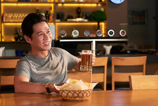 Homem asiático sentado com uma caneca de cerveja e lanches no pub e desviar o olhar para algo