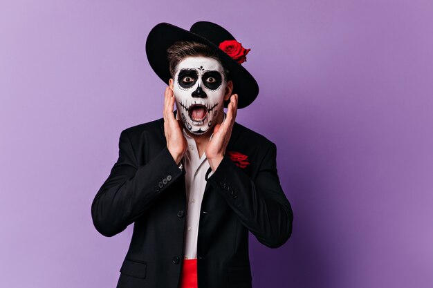 Homem assustado com chapéu de aba larga olhando horrorizado para a câmera. Retrato do cara com maquiagem de Halloween, posando em fundo roxo.