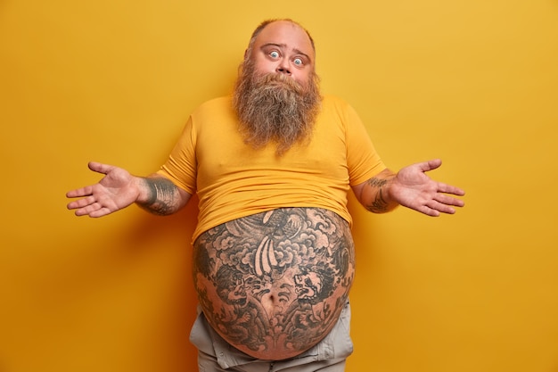 Homem atarracado e hesitante com uma grande barriga tatuada, encolhe os ombros e parece confuso, enfrenta dilemas, toma decisões sérias, veste uma camiseta amarela de tamanho reduzido, posa em ambientes fechados. conceito de pessoas e dúvida