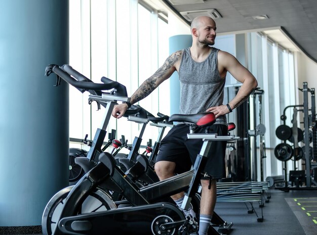 Homem atlético com uma tatuagem na mão ao lado de uma bicicleta ergométrica no moderno clube de fitness