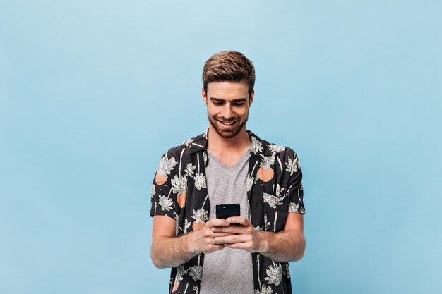 Homem atraente com penteado moderno e barba ruiva em abacaxi e camisa estampada de palma e camiseta cinza sorrindo e segurando o telefone