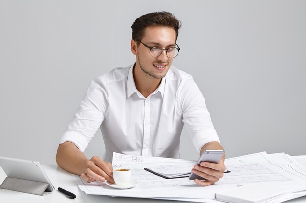 Homem atraente com roupa formal, pausa após trabalho duro, bebe café, digita mensagens no telefone inteligente, tem expressão alegre. Empresário caucasiano usa tecnologias modernas para comunicação