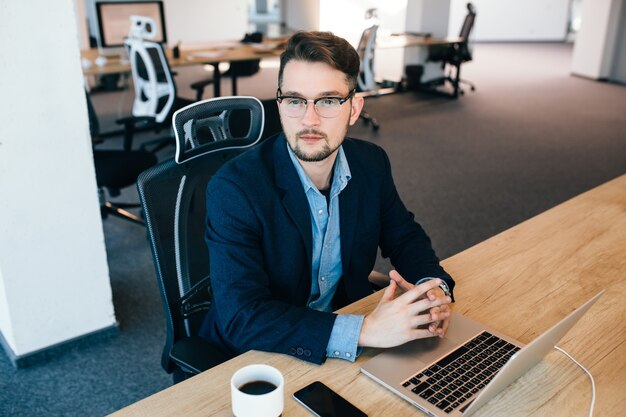 Homem atraente de cabelos escuros está sentado à mesa no escritório. Ele usa camisa azul com jaqueta preta. Ele está olhando para o lado. Vista de cima.