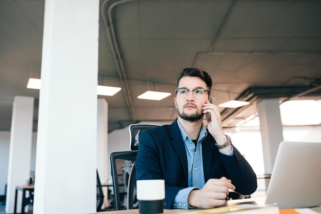 Homem atraente de cabelos escuros está trabalhando na mesa no escritório. Ele usa camisa azul com jaqueta preta. Ele está falando ao telefone.