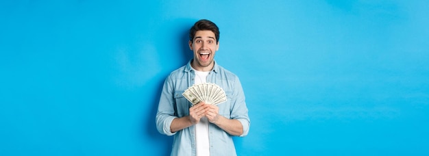 Homem atraente excitado e surpreso segurando o prêmio em dinheiro e sorrindo espantado em pé sobre backg azul