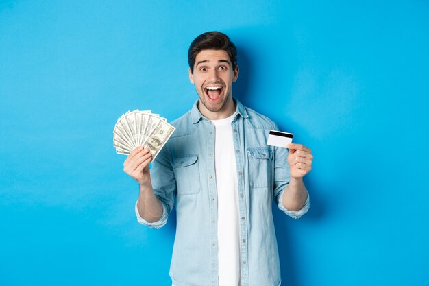 Homem atraente feliz parecendo espantado, mostrando dinheiro e cartão de crédito, conceito de bancos, crédito e finanças. Fundo azul do estúdio