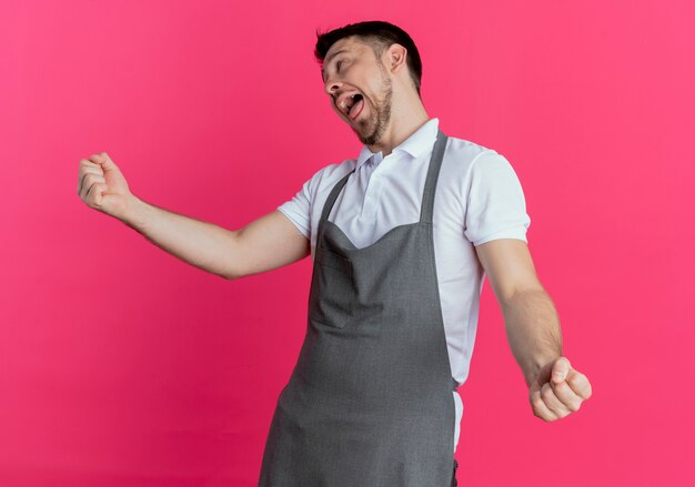 Homem barbeiro com avental louco feliz se divertindo, mostrando a língua em pé sobre um fundo rosa