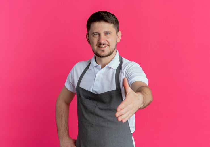 Foto grátis homem barbeiro com avental olhando para a câmera, cumprimentando com a mão e sorrindo amigável em pé sobre fundo rosa