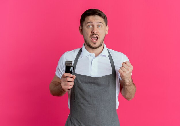 Homem barbeiro com avental segurando o aparador de barba cerrando o punho feliz e animado em pé sobre um fundo rosa