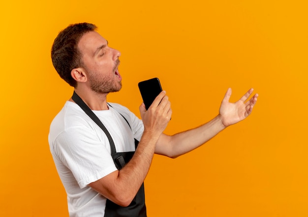 Homem barbeiro com avental segurando um smartphone usando como microfone cantando e se divertindo em pé sobre a parede laranja