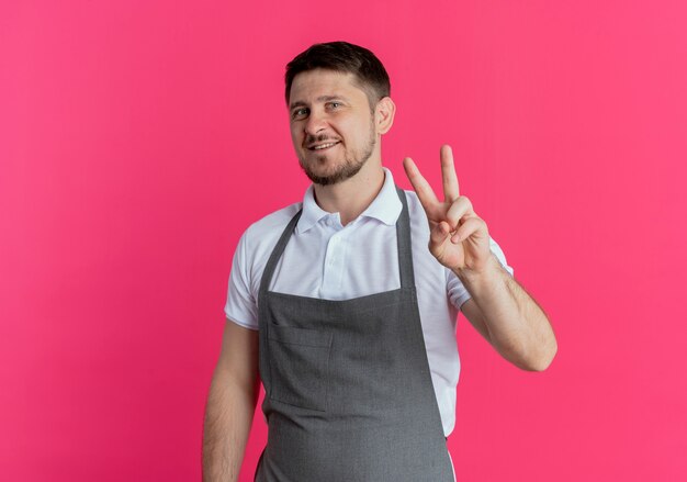 Homem barbeiro com avental sorrindo alegremente para a câmera mostrando o sinal da vitória em pé sobre um fundo rosa