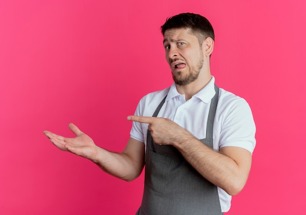 Foto grátis homem barbeiro de avental apresentando com o braço da mão algo apontando com o dedo indicador para o lado em pé sobre a parede rosa